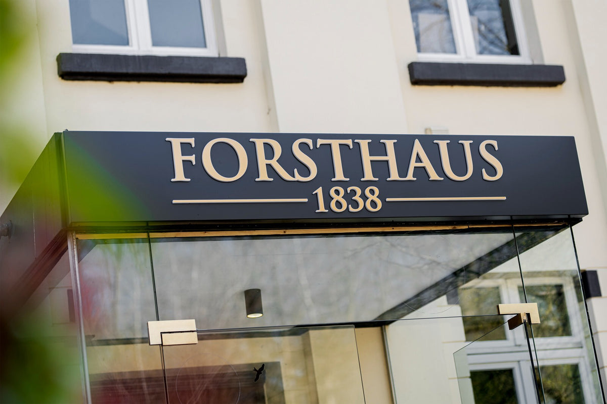 Gläsernes Eingangsvordach mit goldenem Schriftzug Forsthaus 1838 auf dunkler Blende vor heller Hausfassade