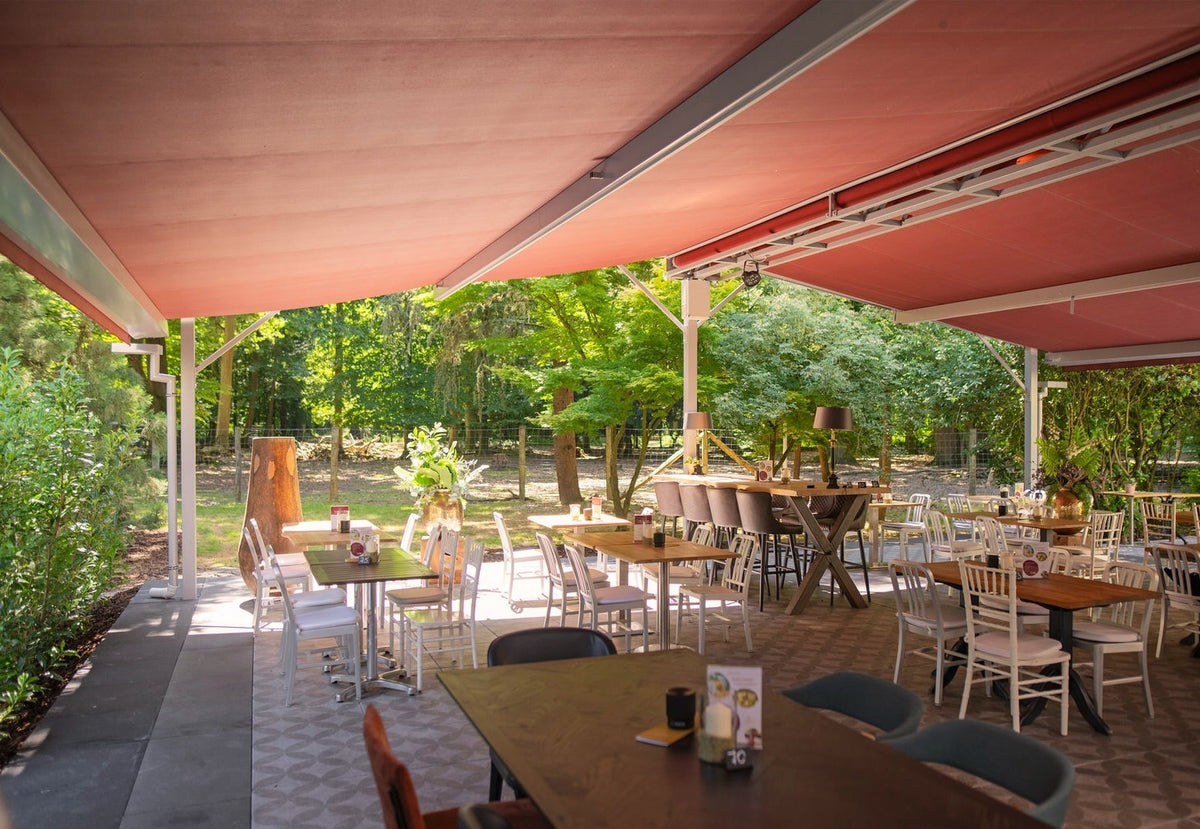 Überdachte Terrasse mit rotbraunen Markisen, Holztischen und weißen Stühlen, dahinter Bäume und ein Zaun