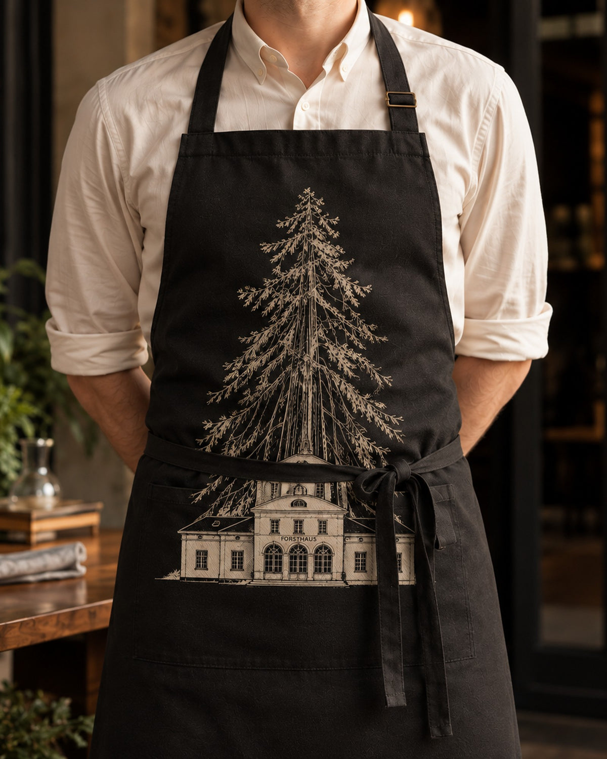 Schwarze Kochschürze Hausrecht, getragen von vorne, mit Baum-und-Haus-Motiv auf der Brust